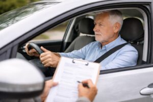 Senior man driving a car during a road test while an examiner in the passenger seat reviews a clipboard in natural daylight.